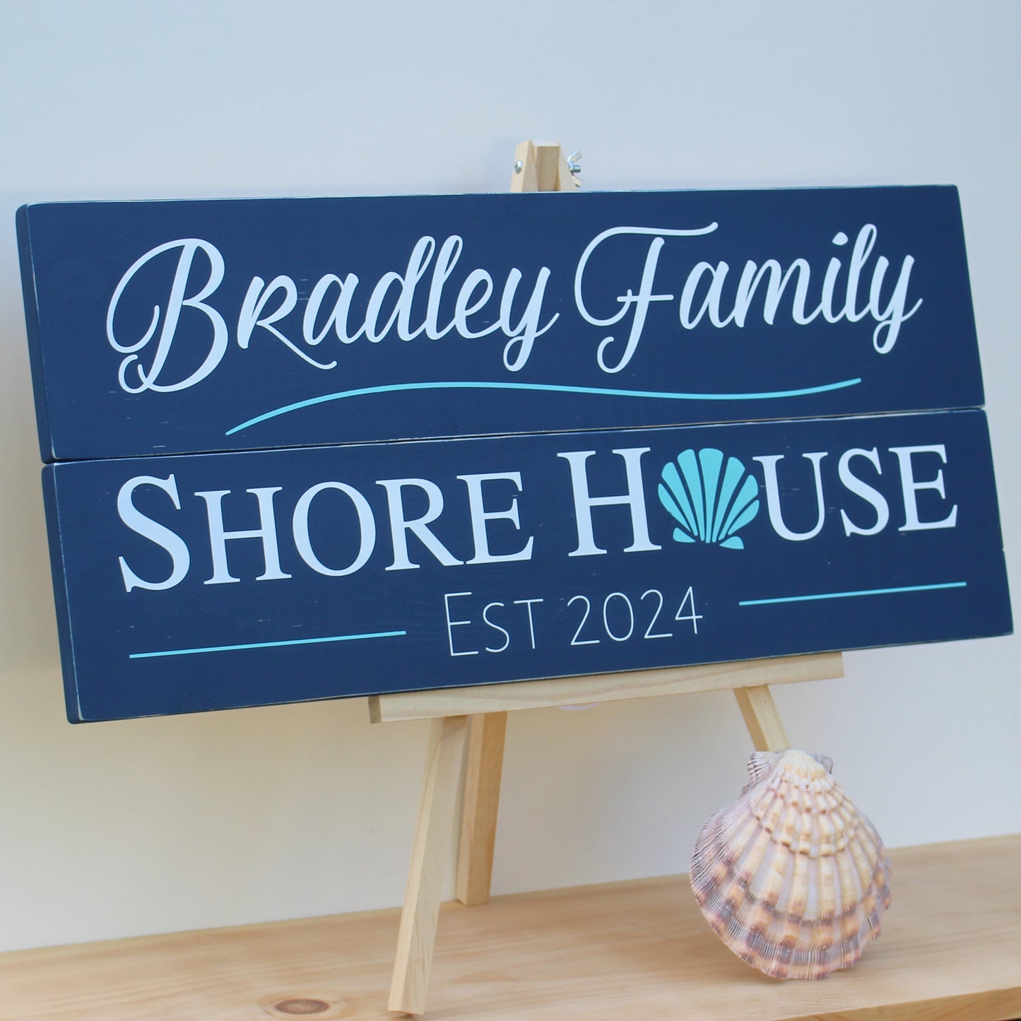 This rustic piece of shore house decor reads, Bradley Family Shore House,Est 2024 and includes a shell graphic. This shore wall art has a navy blue background with white text and aqua graphic details.