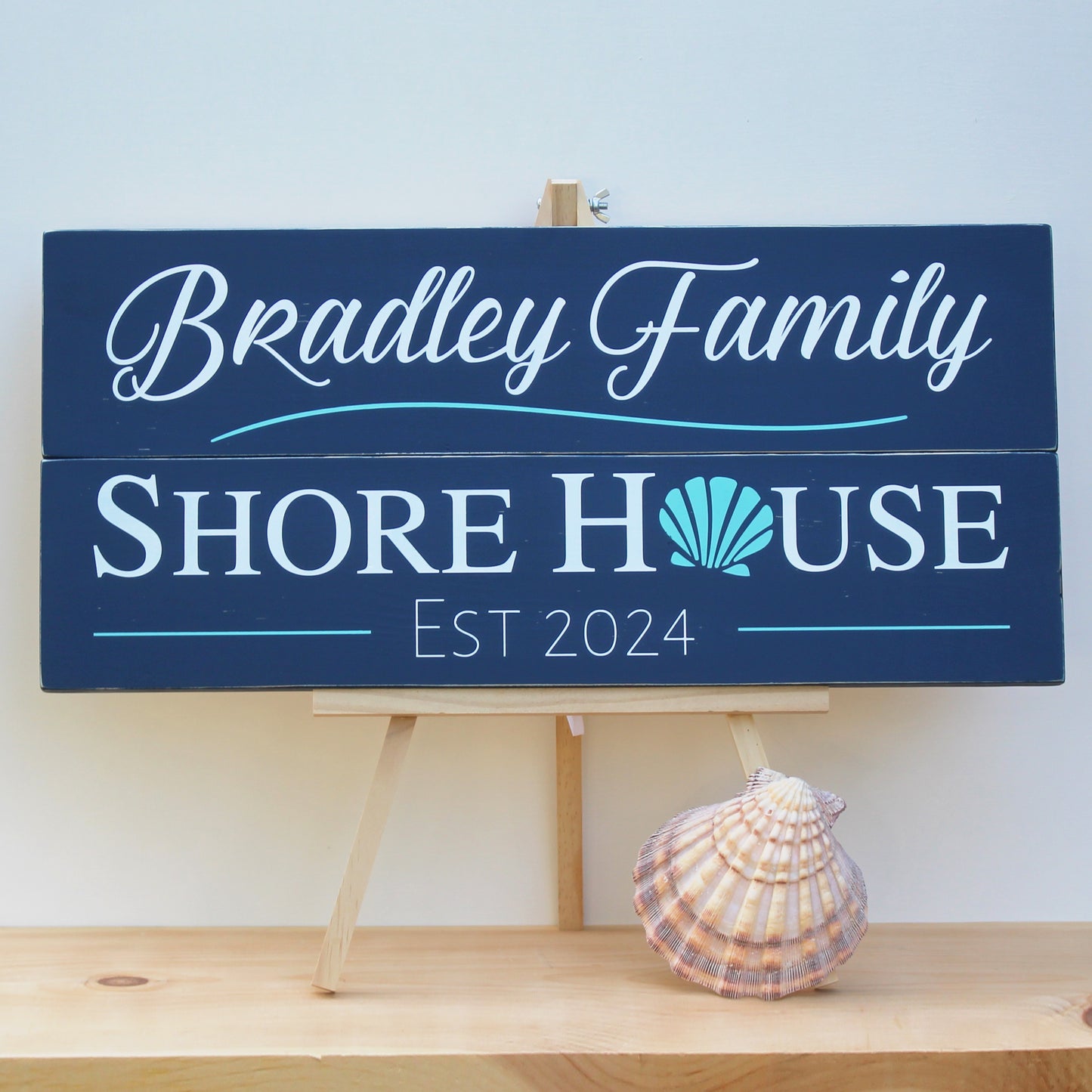 This piece of shore home decor reads, Bradley Family Shore House, est 2024 and features a shell graphic. The background of this shore wall art is navy blue with white text and aqua graphic details.