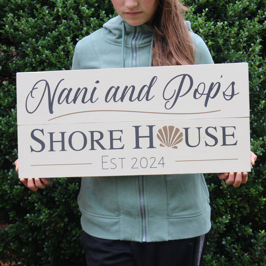 Personalized sea shore wall art that reads, Nani and Pops Shore House, Est 2024.  This coastal farmhouse style sign has a cream background with rustic grey text and muted gold graphics.  The family name and established date are featured.